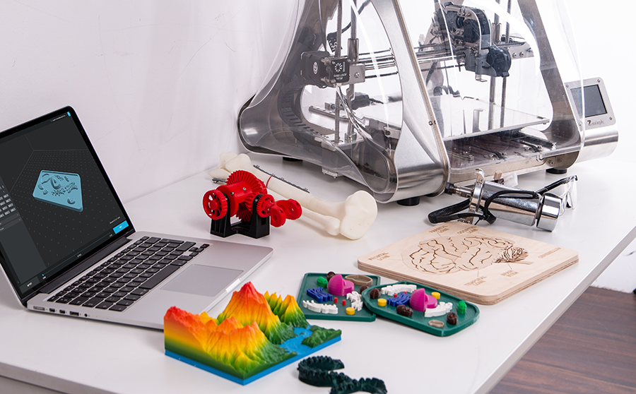 Stock image of a desktop with 3D printed objects, a 3D printer, and a laptop