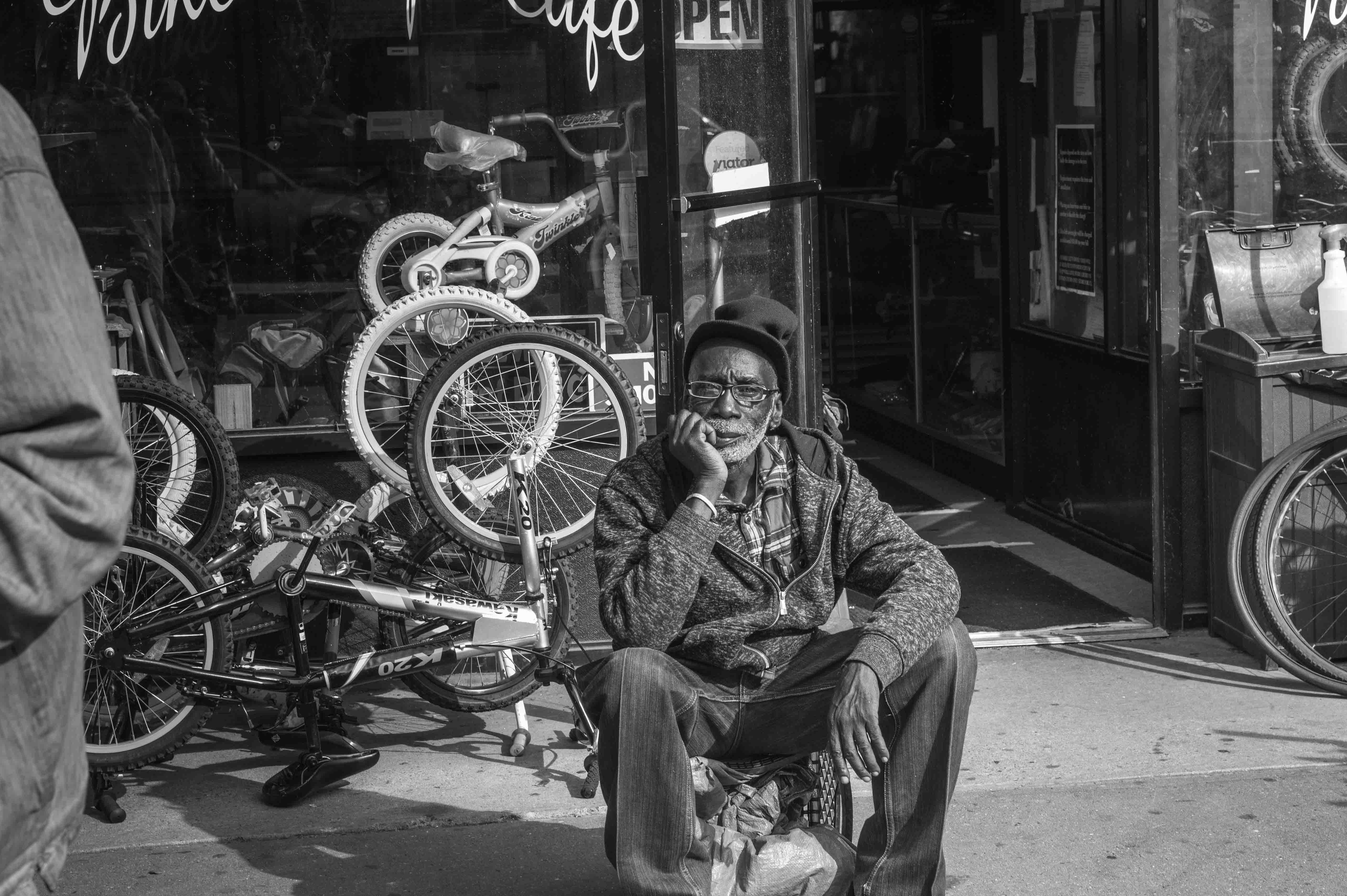Black and white photograph by Hasna Muhammad of an older man sitting on stoop outside of a bicycle shop