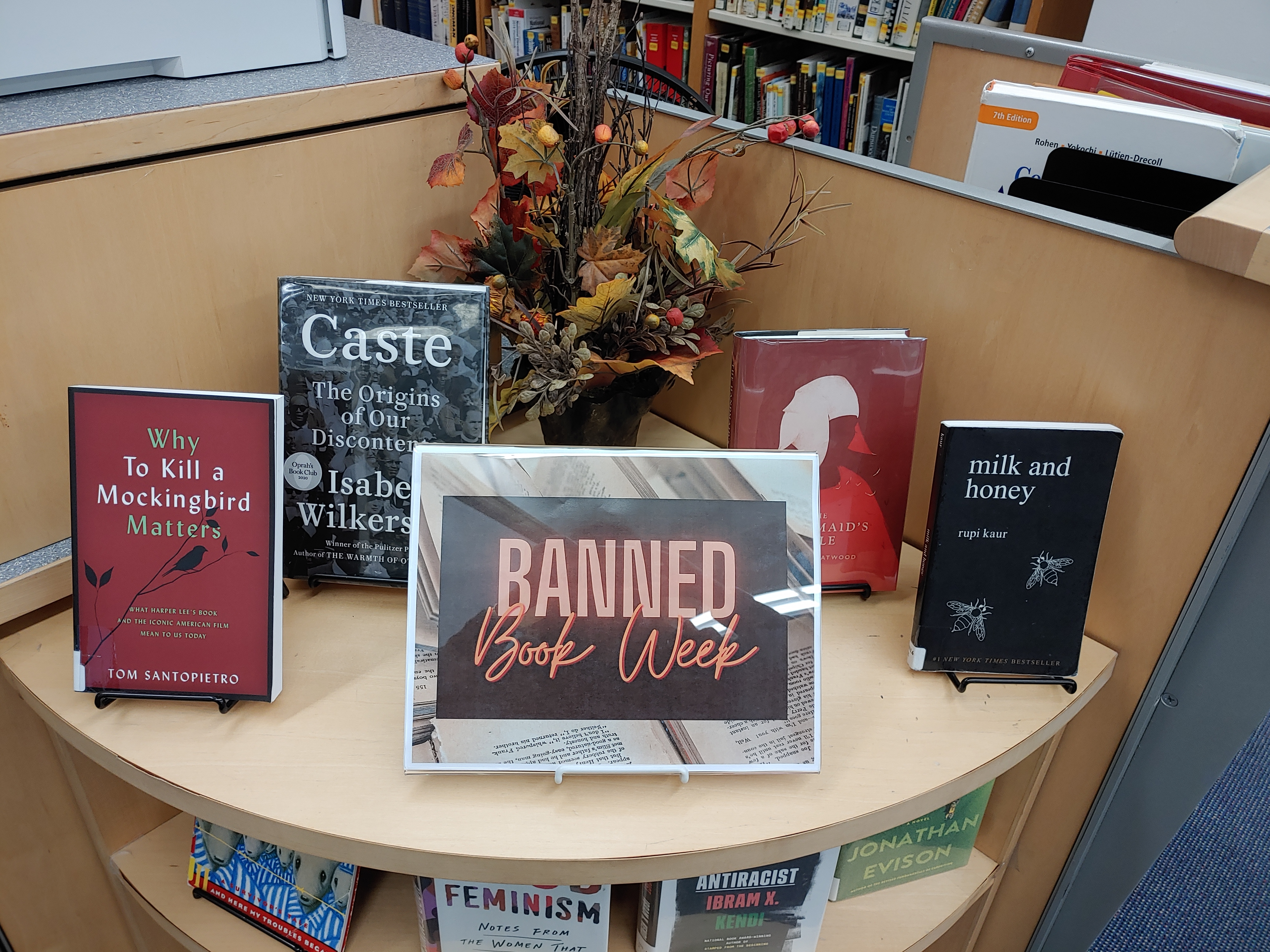 A small bookshelf with four books and a sign reading "Banned Books Week." From left to right, the books are To Kill a Mockingbird, Caste: The Origin of Our Discontents, The Handmaid's Tale, and Milk and Honey, by Rupi Kaur.