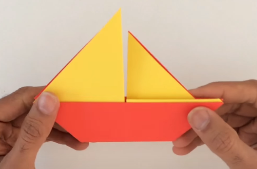Image of an origami sailboat with yellow sails and a red base. It is held in someone's hands on each end.