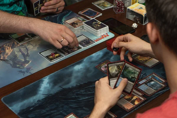 Two players play Magic the Gathering, a trading card game, on a black and blue battle mat; the center of the image is occupied mainly by their hands and cards.