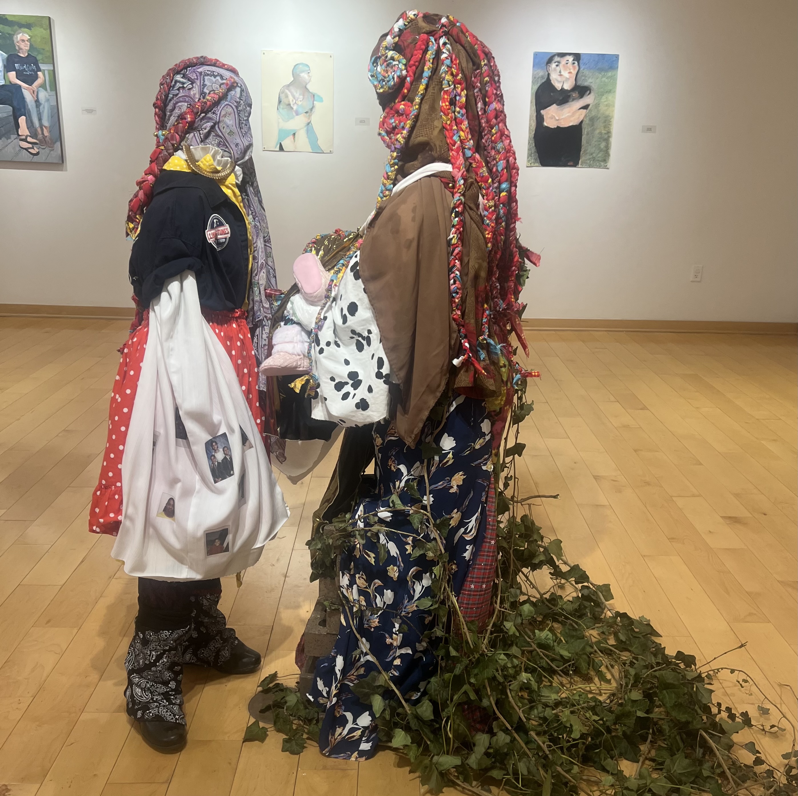 Sculpture depicting two figures facing one another, clothed in multiple kinds and colors of fabric and leaves.