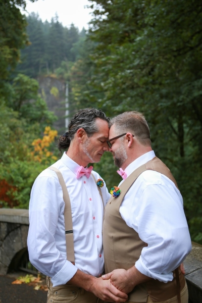 Two men, one in suspenders, the other in a vest, hold hands and press their foreheads together.