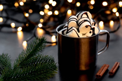 mug of hot cocoa with marshmallows in front of winter twinkle lights and evergreen branch