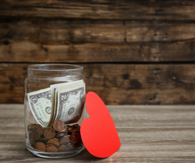 A jar filled with pennies and dollars. A paper cut out of a heart is leaning up against the jar.