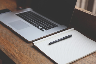 A laptop on a wooden desk. A note book is open next to the laptop, and a pen is resting on top of the notebook.