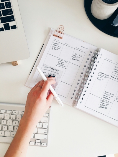 A woman's hand holding a pen and writing in a planner on top of a desk. A keyboard and a laptop are also on the desk.