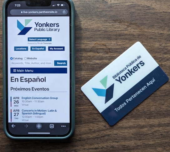 Photo of a smartphone with Spanish language ypl.org on screen and Spanish YPL card "Biblioteca Publica De Yonkers - Todos Pertenecen Aqui"