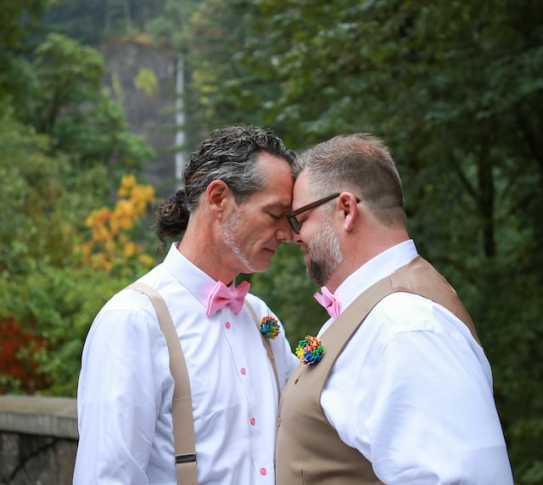 Two men, one in suspenders, the other in a vest, hold hands and press their foreheads together.