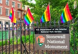 A small sign reading "Stonewall National Monument" on a black iron fence. On top of the fence 4 small rainbow pride flags are stuck on the fence.